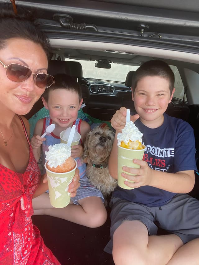 A woman and two children are sitting in the back of a car, enjoying ice cream with whipped cream. A small dog is sitting between the children. The woman is wearing sunglasses and a red outfit, and everyone is smiling