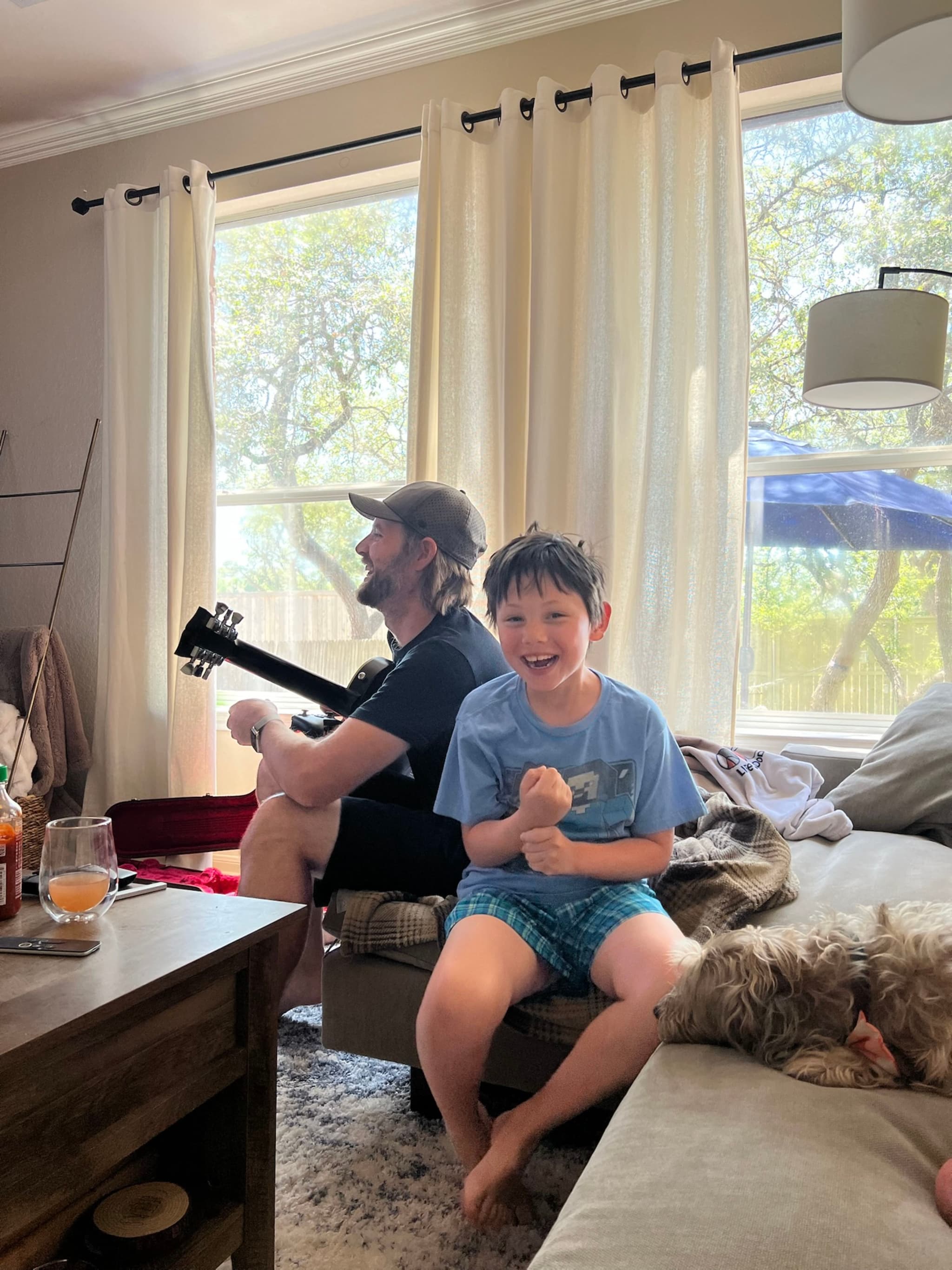 A child sits on a couch smiling, with a person playing guitar in the background. The room is well-lit with natural light coming through large windows, and a dog rests nearby