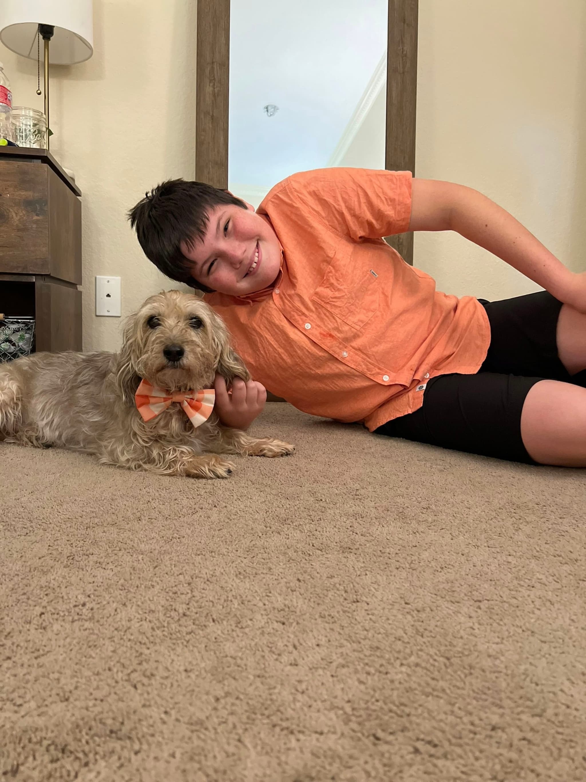 A person lying on the floor next to a small dog wearing a bow tie, both in matching orange outfits, in a room with a mirror and a lamp