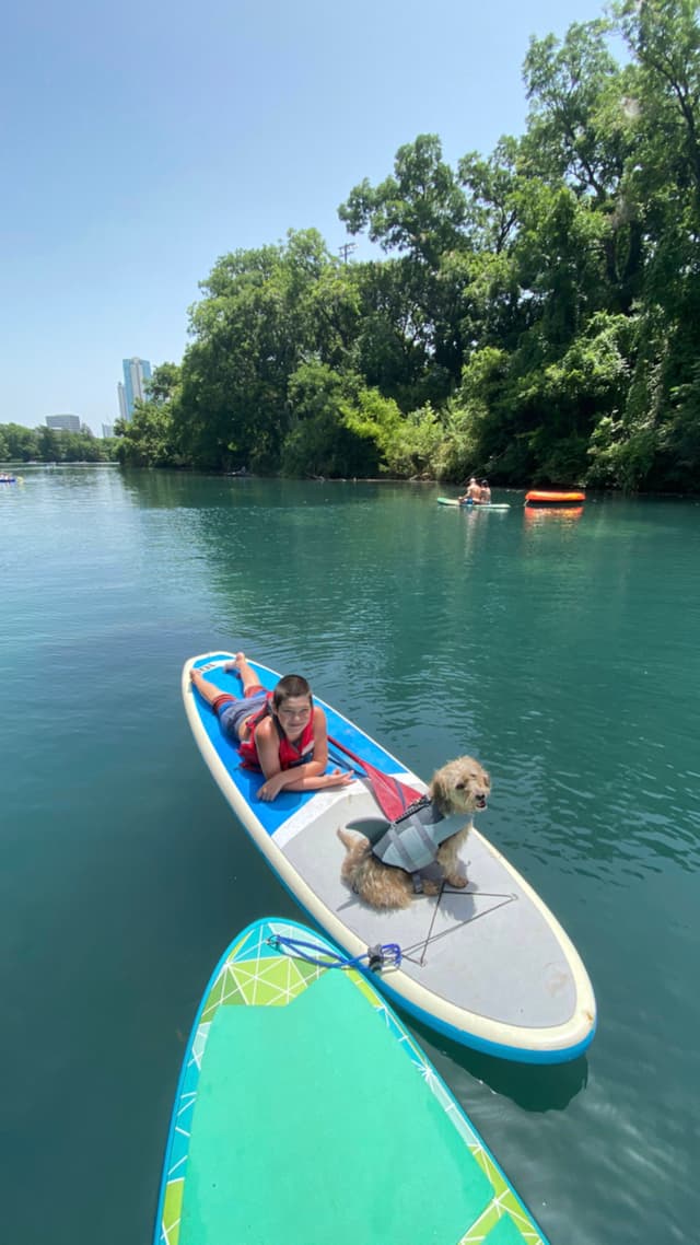 A person and a dog wearing a life jacket are on a paddleboard in a calm, blue lake surrounded by trees, with another paddleboard in the foreground